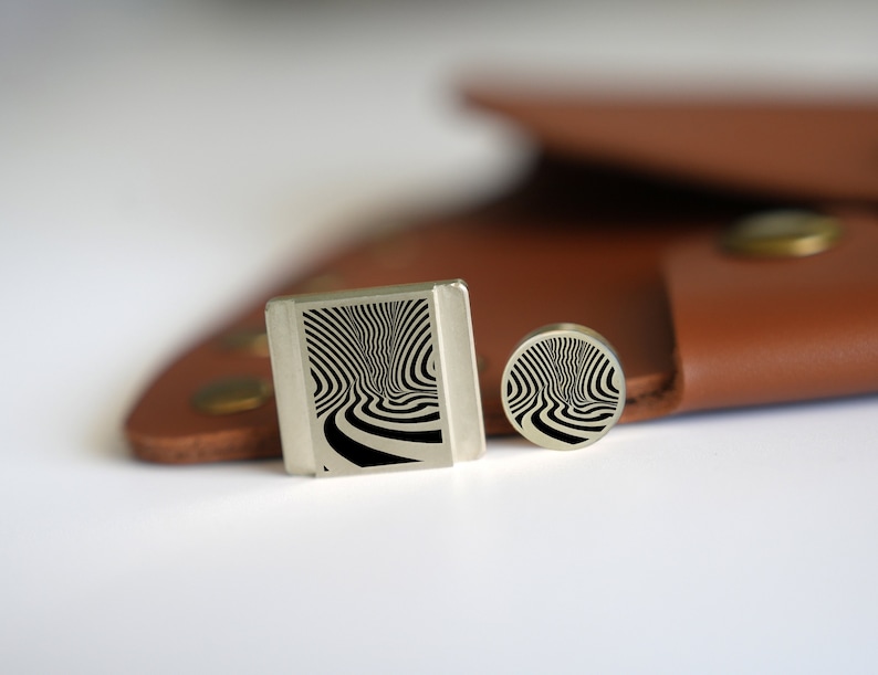 May include: Two silver cufflinks with a black and white swirling pattern. The cufflinks are on a brown leather surface.