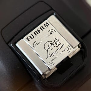 Könnte beinhalten: Nahaufnahme eines schwarzen Blitzschuhs einer Kamera mit einer silberfarbenen Metallplatte. Auf der Platte steht "FUJIFILM" und eine Grafik eines Berges und einer kleinen Figur. Der Text "X-S20" ist ebenfalls sichtbar.