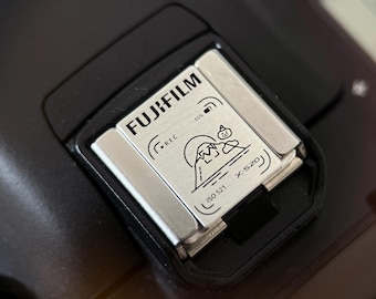 May include: Close-up of a black camera hot shoe with a silver-colored metal plate. The plate has the word "FUJIFILM" printed on it, along with a graphic of a mountain and a small figure. The text "X-S20" is also visible.