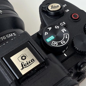 Puede incluir: Primer plano de una cámara Leica negra con una lente plateada y varios diales de control. La cámara presenta el logotipo de Leica y las especificaciones de la lente. El dial de modo está configurado en AUTO. Cámara profesional.