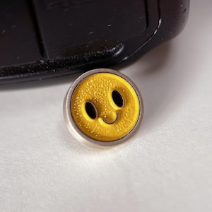 May include: A yellow smiley face button with a textured surface and a silver-colored rim. The button has two oval eyes and a curved smile. The button is on a white surface.