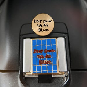 Könnte beinhalten: Eine runde Kamera-Blitzschuhabdeckung aus goldfarbenem Metall mit dem Text "Deep Down We Are Blue." und eine rechteckige Metallabdeckung mit blauem Gittermuster und dem roten Text "Deep Down We Are Blue." mittig auf der Oberfläche.