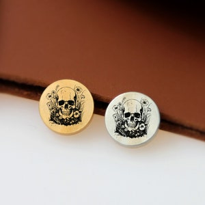May include: Two round metal buttons with a skull and flowers design. The button on the left is gold and the button on the right is silver.