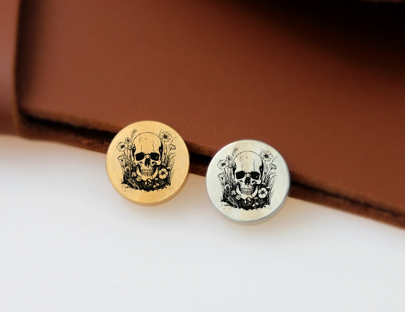 May include: Two round metal buttons with a skull and flowers design. The button on the left is gold and the button on the right is silver.