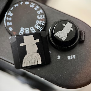 Puede incluir: Primer plano de una cámara negra con un dial plateado y dos accesorios negros. Los accesorios presentan una ilustración estilizada de una persona con sombrero. Un accesorio es cuadrado, el otro redondo.