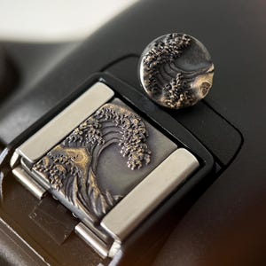 May include: Close-up of a decorative metal camera accessory with a wave design. The accessory is a dark grey colour with silver accents. The wave design is intricately detailed and appears to be made of a dark metal. The accessory is on a black surface.
