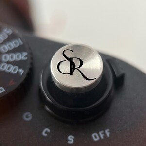 Op de afbeelding: Close-up van een zilverkleurige draaiknop van een camera met de initialen "SR" in zwart schrift. De draaiknop staat op een zwarte camerabody, met andere instellingen zichtbaar op de achtergrond. De camera lijkt een vintage model te zijn.