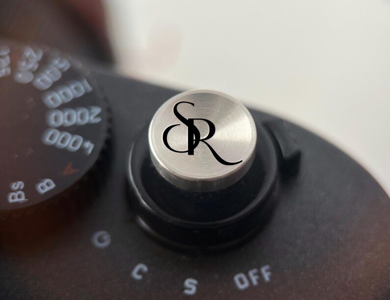 Op de afbeelding: Close-up van een zilverkleurige draaiknop van een camera met de initialen "SR" in zwart schrift. De draaiknop staat op een zwarte camerabody, met andere instellingen zichtbaar op de achtergrond. De camera lijkt een vintage model te zijn.