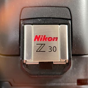 May include: A silver-colored metal hot shoe cover for a camera, featuring the red Nikon logo and the black text Z 30 engraved on the surface. The cover is mounted on the black top housing of a camera body, showing a brushed metallic finish.