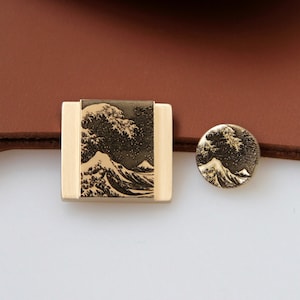 以下が含まれることがあります： 波と富士山が浮き彫りになった、金色の金属製のベルトバックル2つ。バックルは茶色の革の背景の上に置かれています。