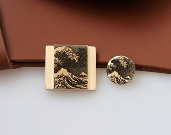 May include: Two gold-toned metal belt buckles with a raised image of a wave and Mount Fuji. The buckles are on a brown leather background.