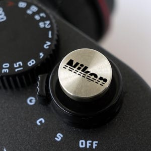 May include: A circular silver-toned metal shutter release button with the black Nikon logo printed on top. The button is attached to a black camera body next to a black shutter speed dial featuring white numbers and a black power switch labeled OFF.