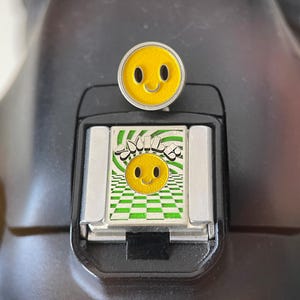 May include: A metal camera hot shoe cover featuring a yellow smiley face with black eyes and a curved mouth. Below it, a square insert displays a green and white checkered pattern, a green swirl background, a yellow smiley face, and the word SMILE.