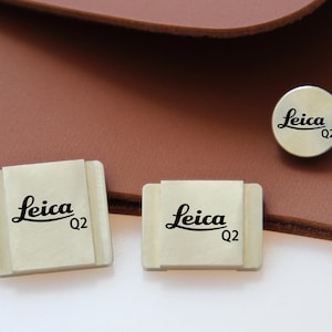Pode incluir: Close-up de acessórios Leica Q2. Dois acessórios retangulares de câmera esbranquiçados com o logotipo Leica e o texto "Q2" são exibidos, juntamente com um acessório redondo. O fundo é uma superfície de couro marrom.