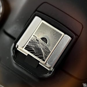 Peut inclure: Gros plan sur le cache griffe d'un appareil photo. Le cache est argenté avec un motif de montagne et de soleil en noir et blanc. Le cache est rectangulaire avec deux pièces latérales argentées. Le corps de l'appareil photo est noir.