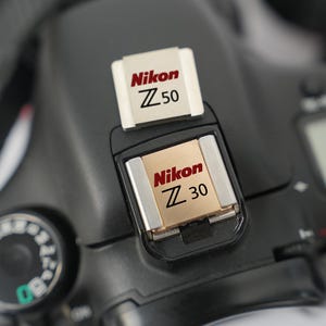 May include: Close-up of a black Nikon camera with two silver and gold flash memory cards. The cards are labeled "Nikon Z 50" and "Nikon Z 30" in red and black text. The camera's dial and strap are visible.