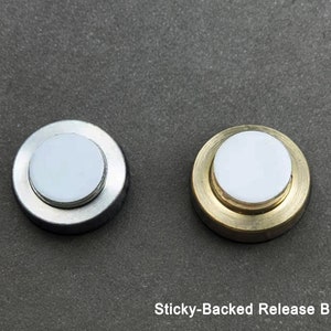 May include: Two sticky-backed release buttons, one with a silver finish and one with a gold finish.  The buttons are round and have a white center.