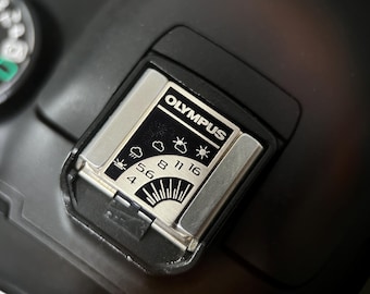 May include: Close-up of an Olympus camera component. The black and silver piece features the Olympus logo and a series of weather-related icons and numbers. The component is set against a black background, highlighting the details of the camera part.