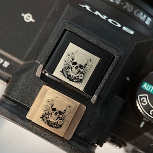May include: A black camera with a silver skull and floral design engraved on a decorative plate. The camera has a dial with the numbers 1, 2, and 3, and the word "AUTO" printed on it.