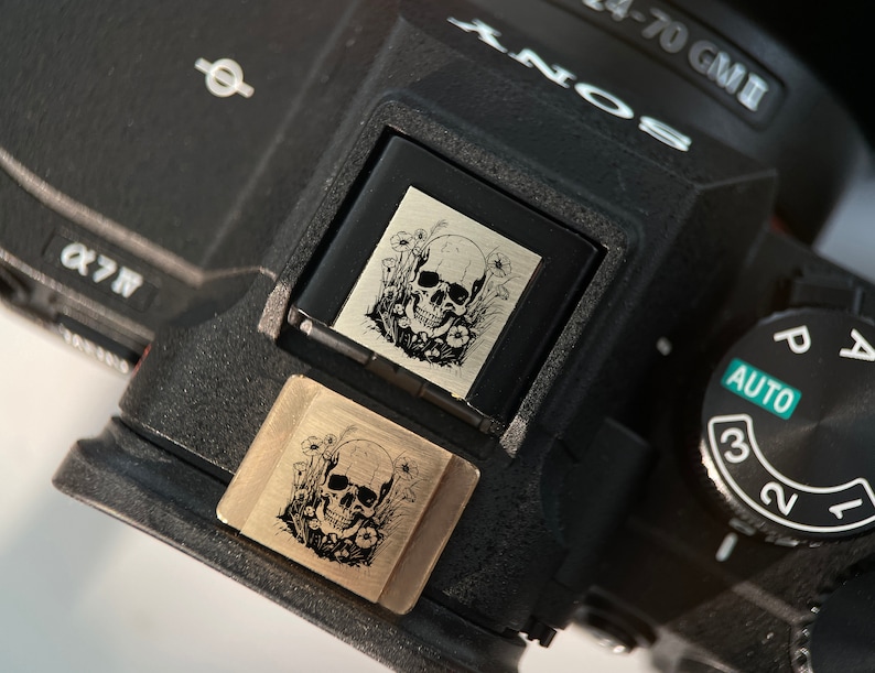 May include: A black camera with a silver skull and floral design engraved on a decorative plate. The camera has a dial with the numbers 1, 2, and 3, and the word "AUTO" printed on it.