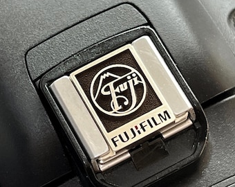 May include: A close-up of a black camera with a silver hot shoe mount. The hot shoe mount has a black plate with the FujiFilm logo and a stylized mountain range.