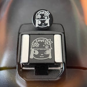 Op de afbeelding: Een zwarte flitsschoenafdekking voor camera's met een witte gravure van een persoon met een helm, het woord LOVE en bloemiconen. Het ontwerp is herhaald op het ronde bovendeel en de vierkante basis die in de camerabevestiging wordt geschoven.