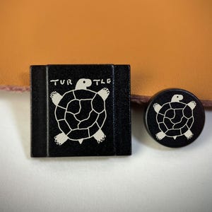 May include: A black square button and a black circular button, both featuring a white line-art illustration of a turtle. The square button has the word TURTLE printed in white above the turtle graphic. Both buttons have a smooth, matte finish.