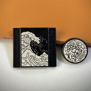 以下が含まれることがあります： 白い波のデザインが施された黒い正方形と丸いオブジェクト。正方形のオブジェクトは長方形で、丸いオブジェクトは円です。波のデザインは、波を様式化した表現です。