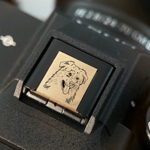 May include: A black camera with a gold-colored metal plate featuring a line drawing of a dog's head. The dog has a black nose, black eyes, and a long tongue sticking out. The camera has a dial with the word "AUTO" and numbers "3" and "2" visible.