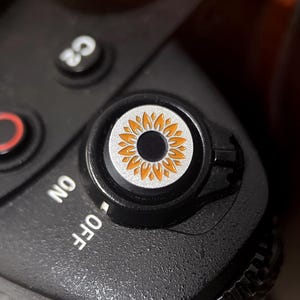 May include: Close-up of a black camera body with a silver and orange sunflower design on a dial. The dial is surrounded by a black ring. The camera has an "ON" and "OFF" switch.