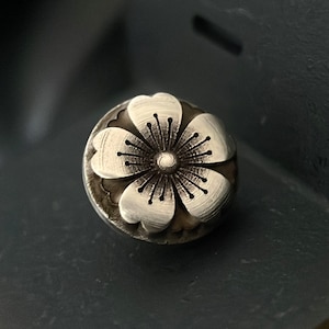 May include: A silver flower charm with a detailed design. The flower has five petals and a center with a small circle.