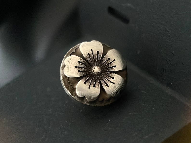 May include: A silver flower charm with a detailed design. The flower has five petals and a center with a small circle.