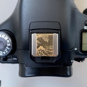 Könnte beinhalten: Eine schwarze Canon DSLR-Kamera mit einer goldenen Blitzschuh-Abdeckung mit einem japanischen Wellenmuster.