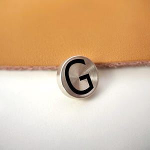 Op de afbeelding: Een kleine, ronde, geborstelde metalen knoop met de zwarte letter "G". De knoop ligt op een wit oppervlak, met een bruin, leerachtig materiaal op de achtergrond.