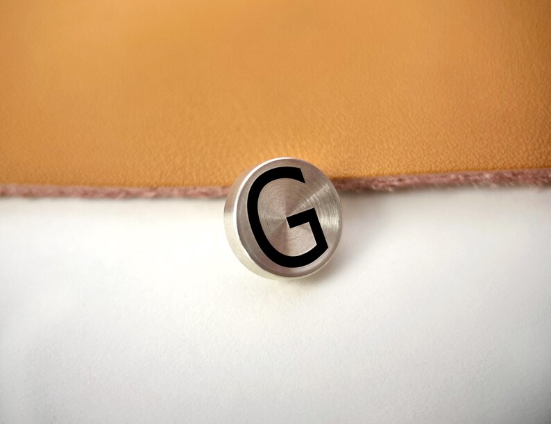 Op de afbeelding: Een kleine, ronde, geborstelde metalen knoop met de zwarte letter "G". De knoop ligt op een wit oppervlak, met een bruin, leerachtig materiaal op de achtergrond.