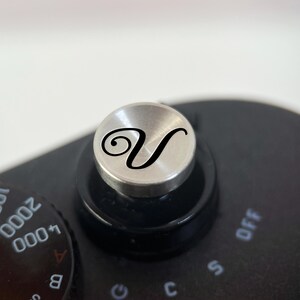 Op de afbeelding: Close-up van de zwarte behuizing van een camera met een zilveren draaiknop met een zwarte cursieve letter "V". De draaiknop is omgeven door een zwarte ring en zit bovenop de camerabody. Een zwarte draaiknop met witte cijfers en letters is zichtbaar.