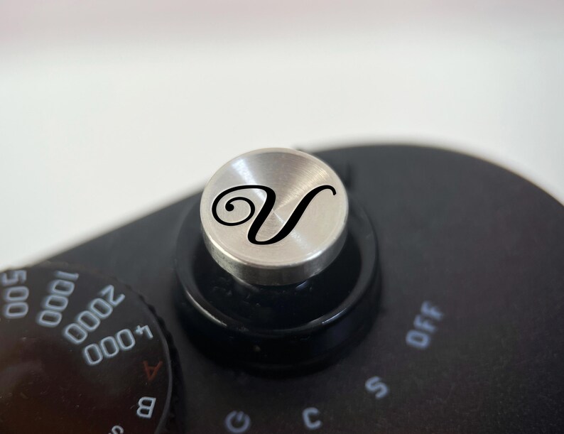 Op de afbeelding: Close-up van de zwarte behuizing van een camera met een zilveren draaiknop met een zwarte cursieve letter "V". De draaiknop is omgeven door een zwarte ring en zit bovenop de camerabody. Een zwarte draaiknop met witte cijfers en letters is zichtbaar.