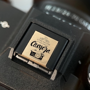 May include: Close-up of a black camera with a gold-colored accessory. The accessory has the text "You don't need a time machine if you have a camera" with a camera illustration. The camera has an auto setting.