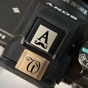May include: A black camera with a silver and black custom plate that says "A" with a mustache above it. The camera has a silver plate that says "W" on it.