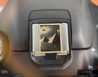 May include: A rectangular camera hot shoe cover featuring a gold-toned metal frame and a black inlay. The inlay displays a detailed engraved graphic of two pyramids under a swirling galaxy and a crescent moon, set against a starry night sky.