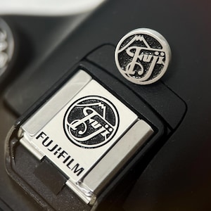 Op de afbeelding: Close-up van een zwarte camera met een zilveren Fujifilm-logo op de flitsschoen. Een zilveren speld met het Fujifilm-logo en een bergontwerp ligt erbovenop. De draaiknop van de camera is zichtbaar op de achtergrond.