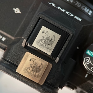 May include: A black camera with a gold and silver decorative plate featuring a skeleton riding a flamingo. The plate is attached to the top of the camera. The camera has a dial with the numbers 1, 2, and 3, and the word "AUTO" printed on it.