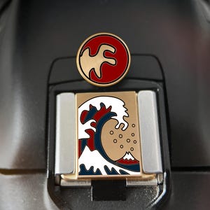 May include: A circular enamel pin featuring a red center with a gold abstract shape, positioned above a rectangular hot shoe cover. The cover displays a graphic of a large white wave with blue and red accents and a mountain peak on a gold background.