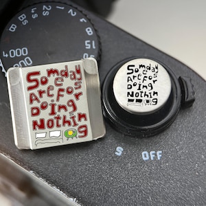 Op de afbeelding: Een vierkante metalen flitsschoenafdekking en een ronde ontspanknop voor camera's, beide voorzien van de tekst "Someday Are For Doing Nothing" met een kleine afbeelding van een bed en een koffiemok. De vierkante afdekking heeft rode letters, de knop zwarte.