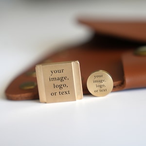 Op de afbeelding: Twee goudkleurige metalen stempels met de tekst "your image, logo, or text" erop gegraveerd. De stempels liggen op een bruin lederen oppervlak.