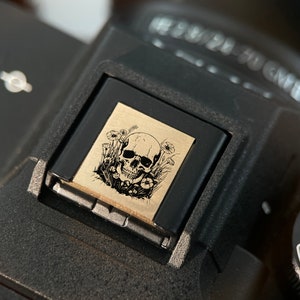 May include: A black camera with a skull and flowers design on the hot shoe mount. The camera has a dial with the word "AUTO" and numbers "3" and "2" visible.