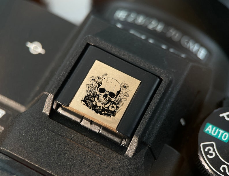 May include: A black camera with a skull and flowers design on the hot shoe mount. The camera has a dial with the word "AUTO" and numbers "3" and "2" visible.