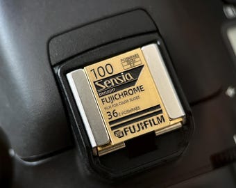 May include: Close-up of a black camera with a gold and silver film cartridge. The cartridge reads "100 Sensia Daylight Fujichrome" and "36 E-POSWFAE". The Fujifilm logo is visible.