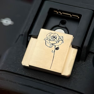 May include: A gold-colored camera hot shoe cover with a black line drawing of a rose.