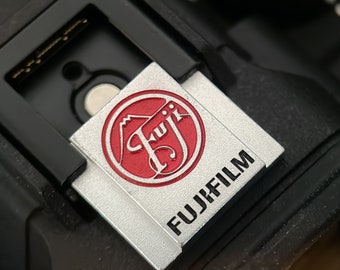 May include: Close-up of a camera accessory with the Fujifilm logo. The accessory is silver with a red circular emblem. The Fujifilm text is in black. The accessory is attached to a black camera body.
